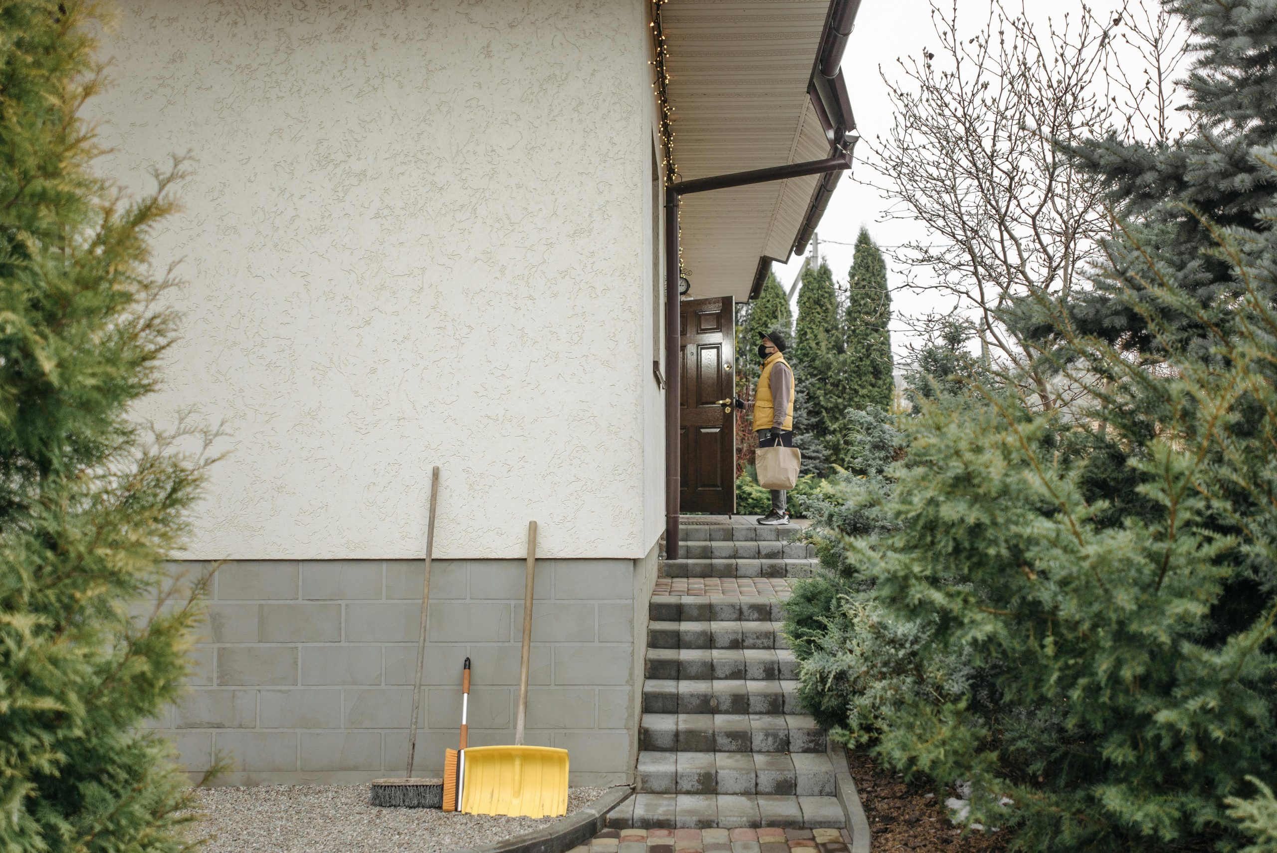 Delivery person holding a paper bag standing at a house door, surrounded by outdoor greenery.
